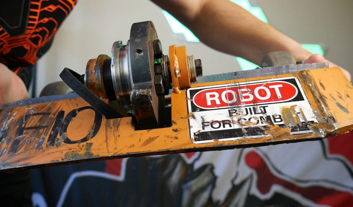 Battle damage! Angus Deveson, post-competition inspection: Hands holding up a large metal damaged looking combat robot (beetle-weight class). Scratches and gashes are evident on the robot's front, the thick metal spinner blade is blunted at the end.