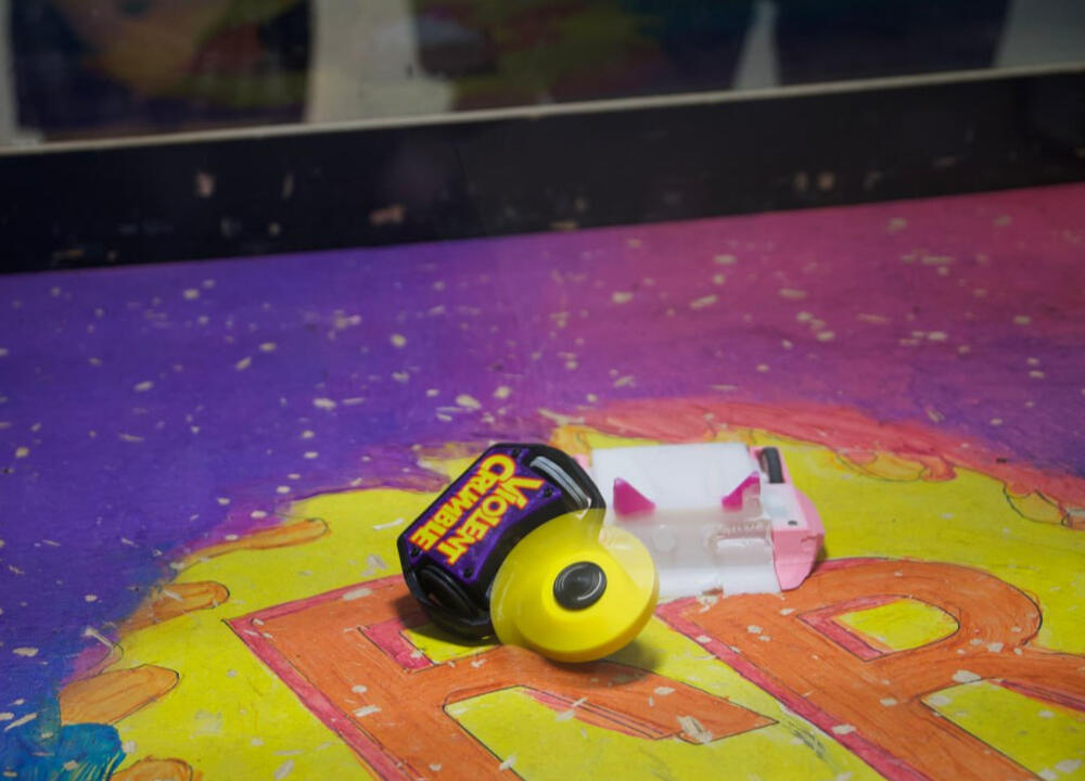 A close up in an area, where small ant-weight class robots are battling, primarily 3D Printed. Violent Crumble (purple robot with a candy wrapping looking title, with a thick yellow plastic blade spinning like crazy), taking on another robot that is a ramp