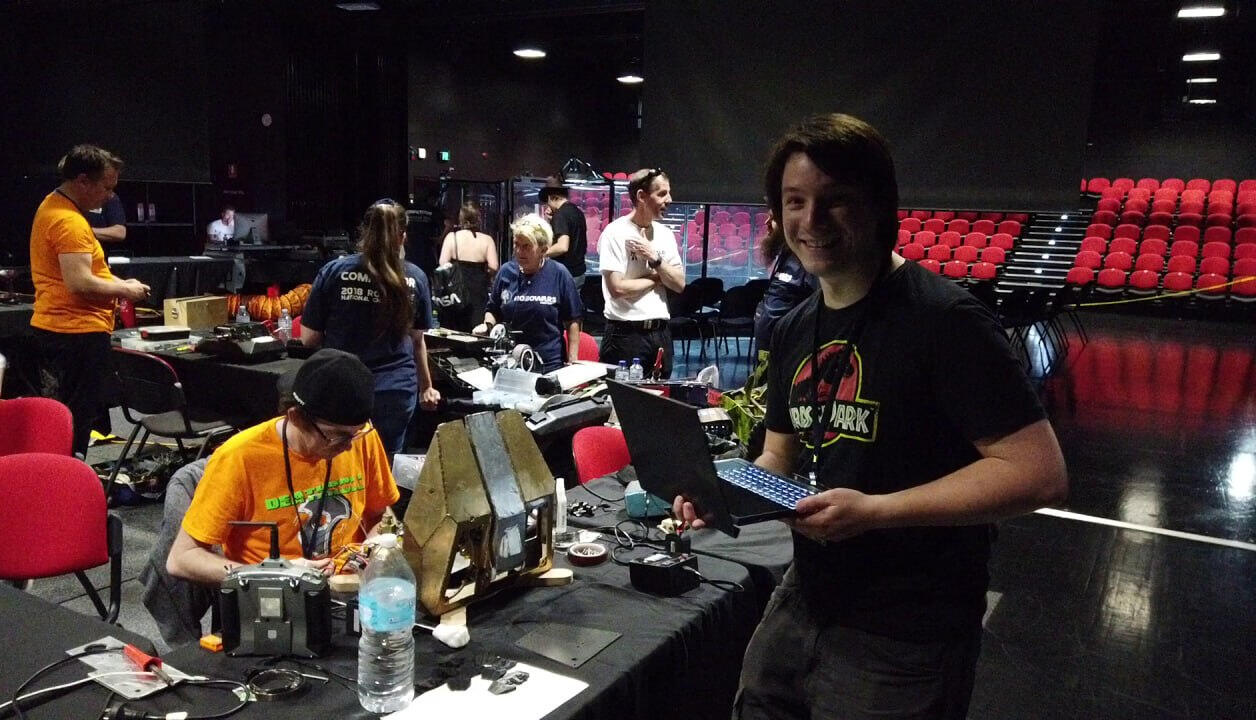 Angus Deveson at a past Robot Wars Competition in Australia: Standing to the side next to the competitors work stations while they set up and tend to their robots, smiling and dressed in a Jurassic Park black shirt, holding a laptop.