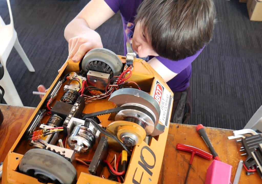 Angus Deveson at a robot event: close up shot of Angus peering into a large metal combat robot (Beetle-weight class), examining the wheels. Predominantly metal with a tidal wave-looking thick spinner blade weapon.