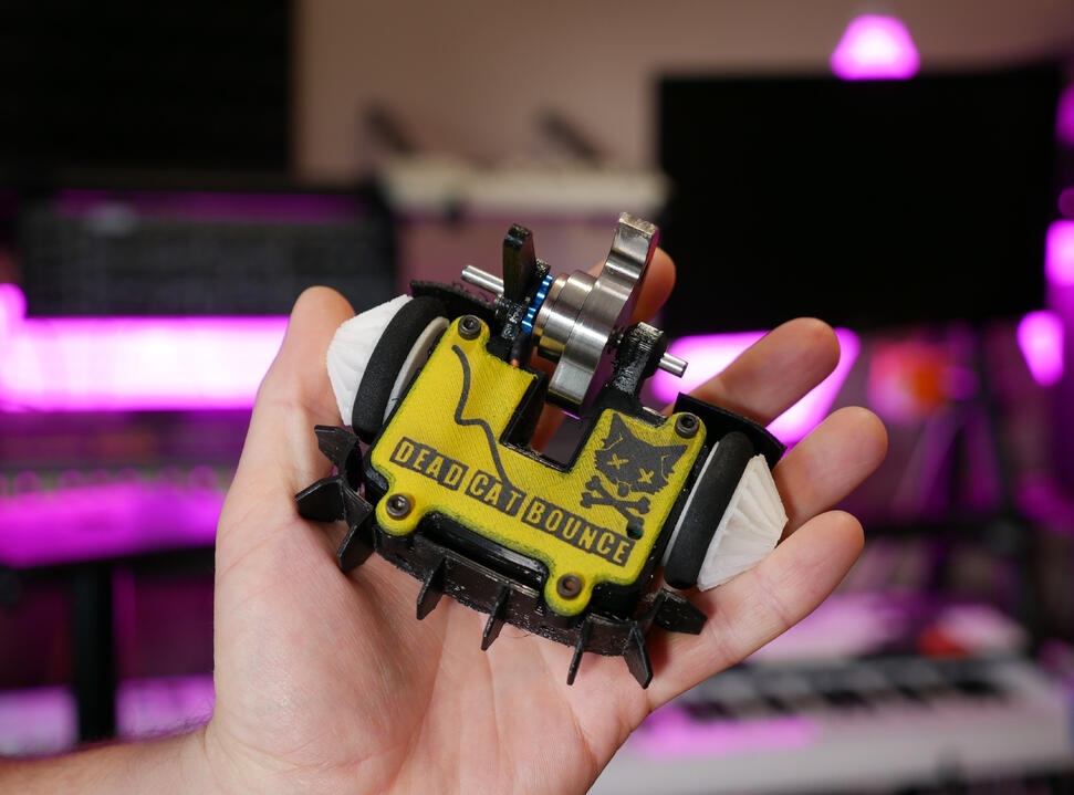 Angus's hand holding up small combat robot in his studio: Ant-weight class, with a metal spinner blade custom made. 3D printed yellow cover with a black logo "Dead Cat Bounce" on it. Has a graphic design of a goofy black cat with "x" eyes and cross bones.