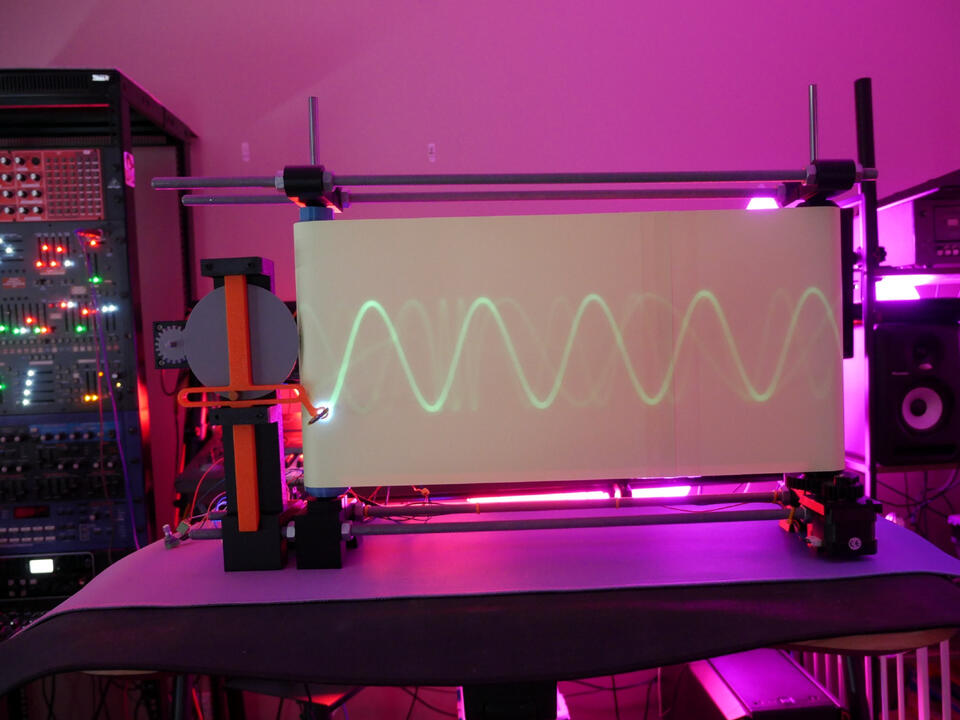 Scotch Yoke Visualizer A machine showing a visual pathway of the scotch yoke mechanism working: using green LED tracing on a rotating parchment slab, the machine set on a table in a workshop, bathed in pink lights.