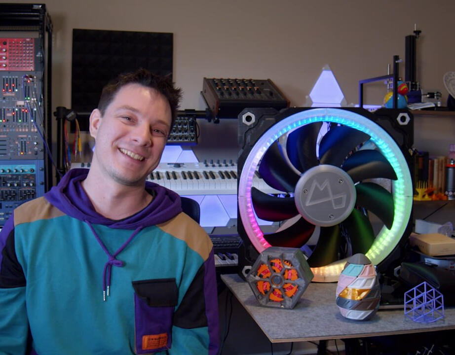 Angus Deveson in his workshop, standing beside his 3D printed projects which is on a table to the right. Featuring: a giant 3D printed RGB fan, a 3D printed Makers muse puzzle box, and a 3D printed puzzle box that looks like an egg.