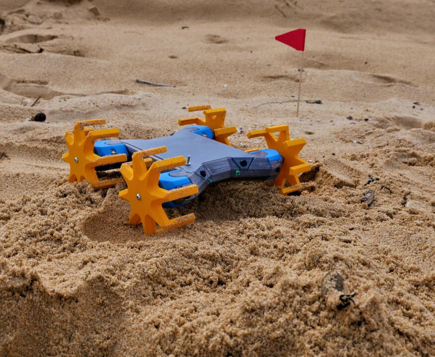 Makers Muse's Off-Roader Vehicle, primarily 3D Printed! It's a small blue robot/vehicle with 4 spindly looking orange wheels, the vehicle navigating over the beach sand around a tiny red flag sticking out of the sand.