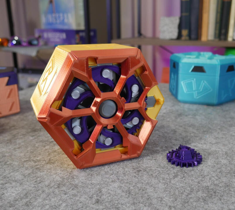 A close up look at Makers Muse's 3D printed expanding puzzle-box design on a gray surface: Hexagon shaped and coloured orange and purple. Many mechanisms and gears are evident in the design.